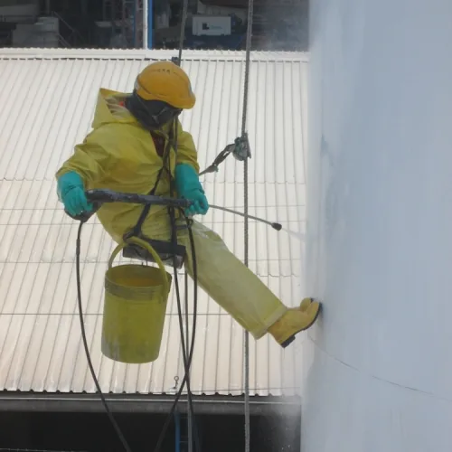Operario de GEINCOR suspendido en un arnés de trabajo con dos cuerdas, traje amarillo, casco, protección facial y guantes, lavando la pared exterior de un tanque.